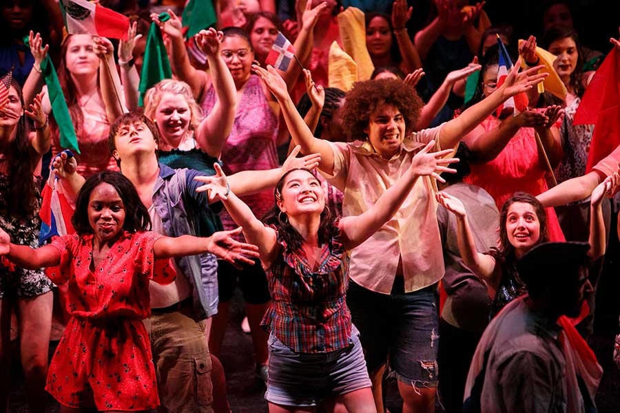 A large crowd of Muse student cast members look and reach upward toward the balcony